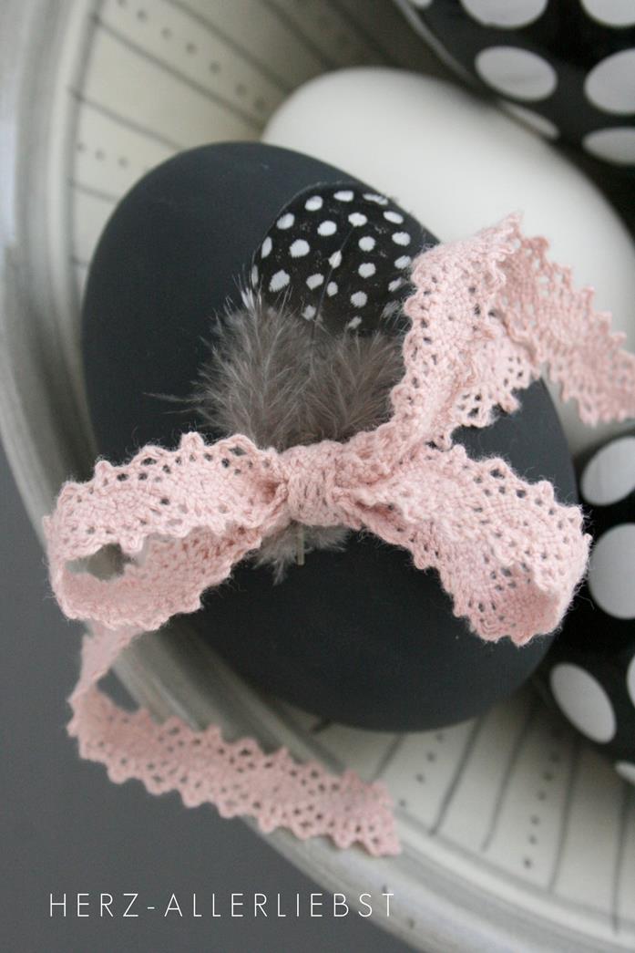 dark grey egg, dyed with matte paint, decorated with pale pastel pink ribbon, made from pink crochet lace, and a small quail feather, easter egg decorating, placed in dish, near other black and white eggs