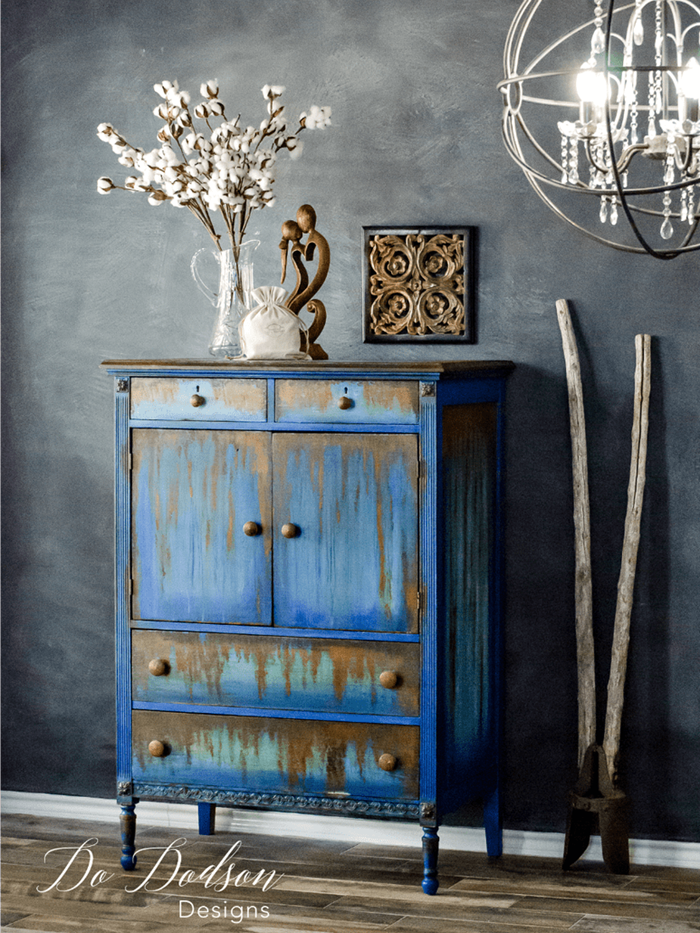 bright blue vintage cabinet, with orange paint effect resembling rust, near dark grey unevenly painted wall, and various decorations