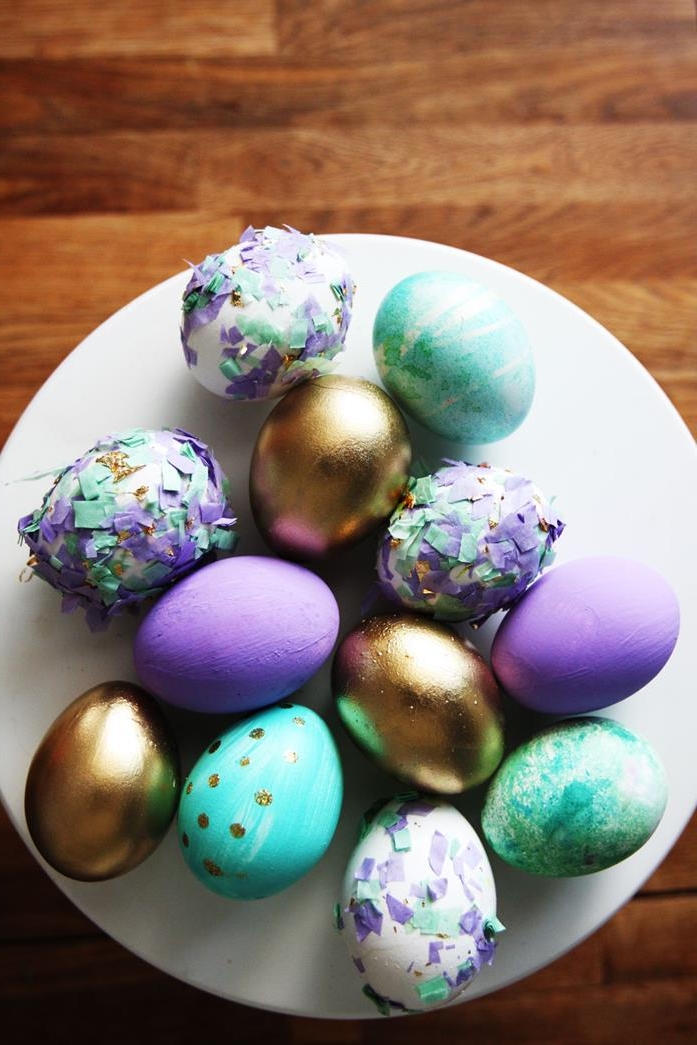 dying easter eggs, round white dish, containing eggs dyed in purple, turquoise and shiny golden color, some have patterns, while others are decorated with colorful broken egg shells