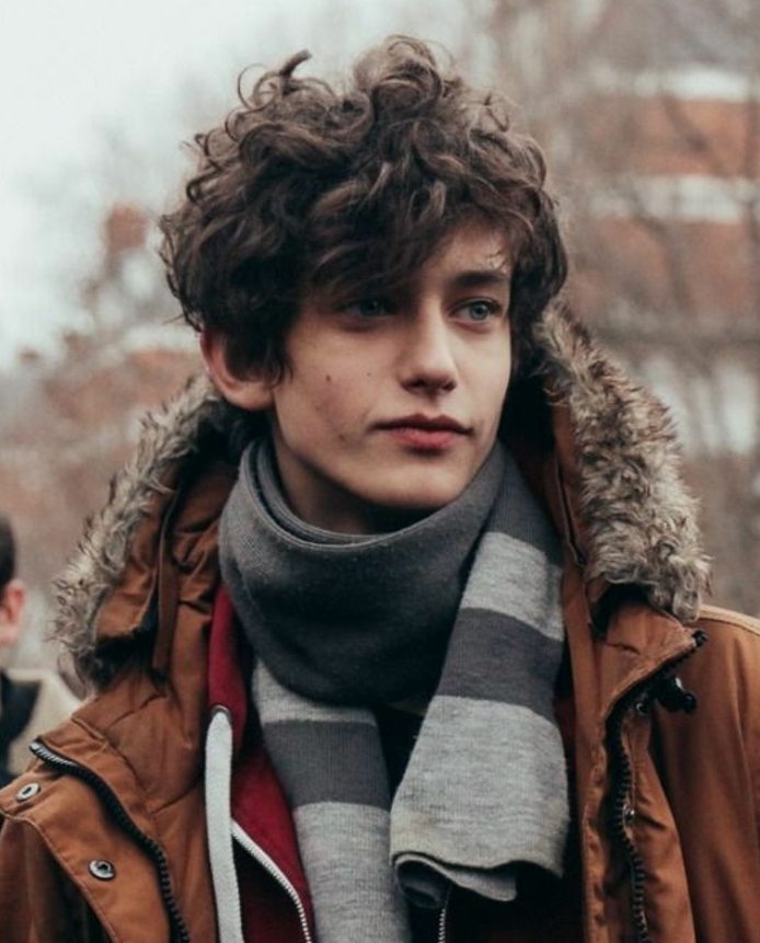 smiling teen with brunette, curly and messy hair, hair designs for boys, rebellious look, combined with brown, hooded winter jacket, and grey striped scarf