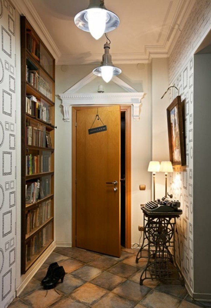 ajar wooden door, leading into a hall, with floors covered in rough stone tiles, patterned wallpaper and wooden bookshelves, antique typewriter on table, near two lamps and a framed painting, hallway decor ideas