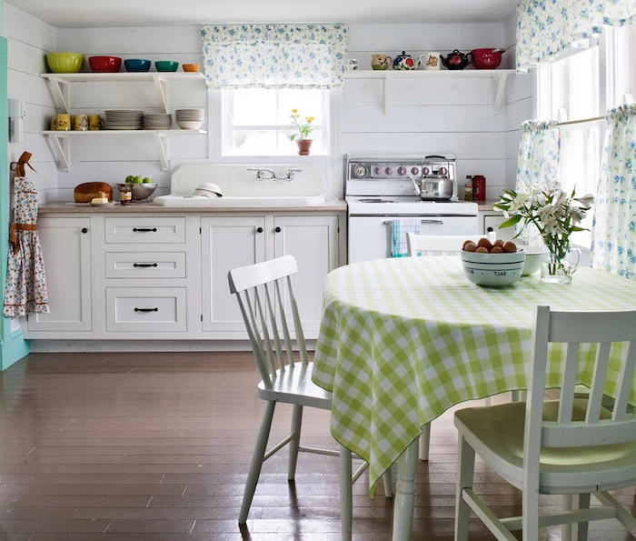 shabby chic decorating, round table with light green and white chequered tablecloth, two mismatched white chairs, brown wooden floor, white kitchen cupboards, and a vintage stove