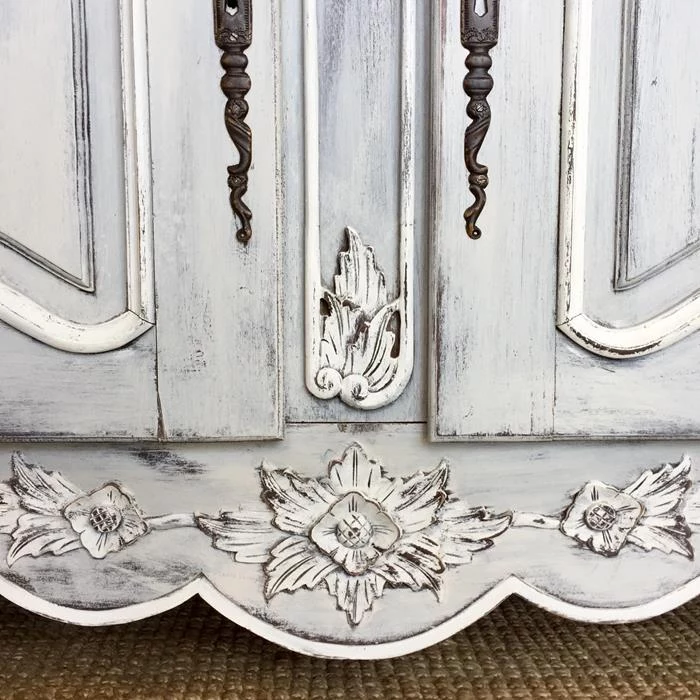 close up of an engraved wooden detail, on antique cupboard, unevenly painted in off-white color, ornamental metal handles