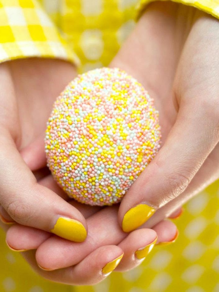 sprinkle-covered ester egg, in yellow and white, pale blue and pink, held by two hands with yellow nail polish, and white and yellow chequered sleeves