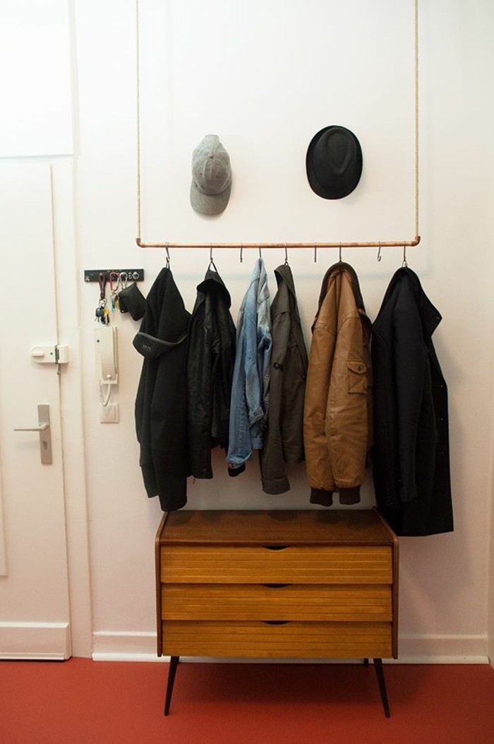 minimalist clothes hanger, hanging from ceiling, with several coats hung on it, brown retro cupboard underneath, red floor and white walls, hallway design ideas, two hats hung on wall