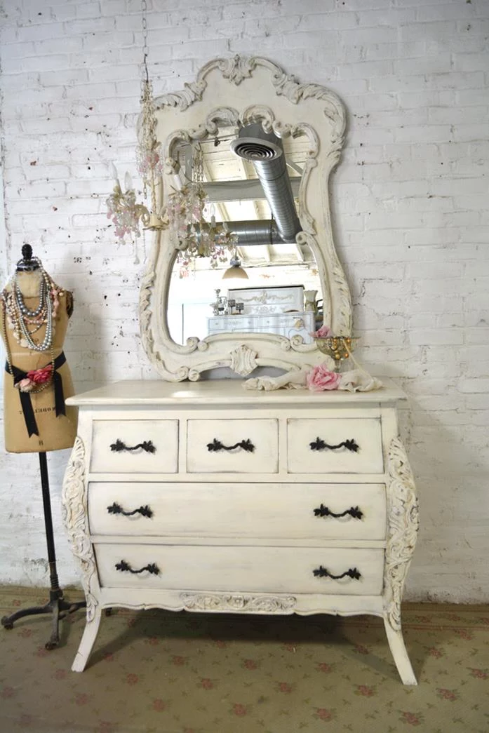 faded floral carpet, under a cream-colored dresser, with ornamental mirror, and black wrought-iron handles, near dressmaker's dummy
