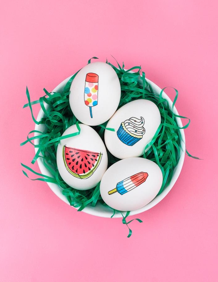 watermelon slice, ice lollies and cupcake stickers, on white hen's eggs, placed in a white bowl full of green easter grass, pink background