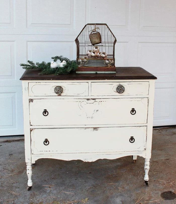 birdcage with vintage look, containing cristmas baubles, country chic décor, placed near a pine branch, on an off-white distressed cupboard, with brown top
