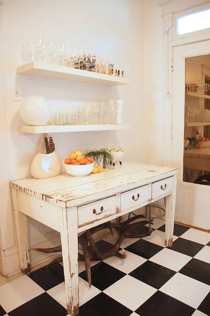 shelves made of wood and painted white, over an antique shabby chic kitchen table, with peeling white paint, black and white tiled floor, glasses and decorations