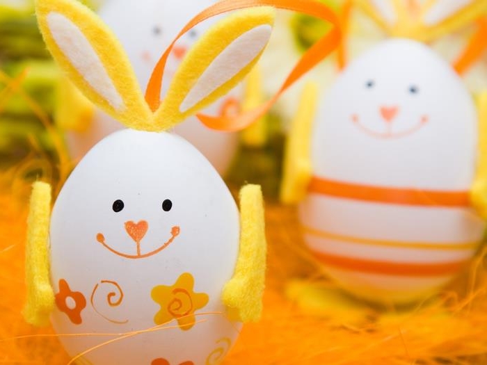 bunny made from white egg, decorated with a hand-drawn face, yellow felt ears and arms, and small flower stickers, easter egg designs, more similar eggs in the background