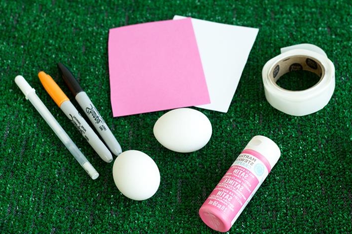 sheets of paper in white and pink, placed on artificial grass, near roll of white masking tape, a tub of pink paint, two white eggs, and three markers, coloring easter eggs 