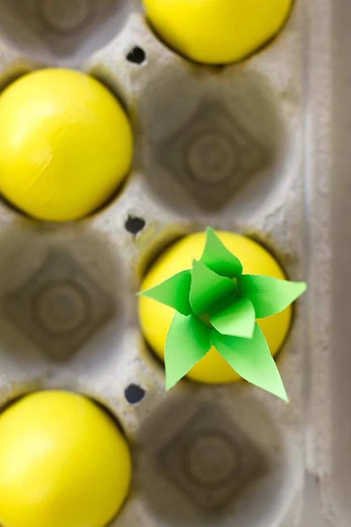 close up of four eggs inside a cardboard box, all painted in yellow, one of them has several paper leaves stuck to its top, making it look like a pineapple, dying easter eggs 