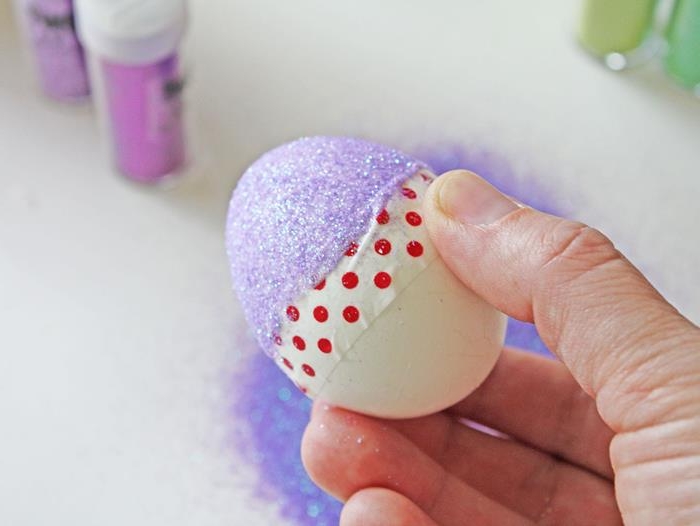violet glitter covering half of an egg, the other half is left white, there is washi tape stuck across the middle, coloring easter eggs, a hand is holding the egg