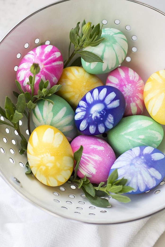 violet and pink, yellow and green dyed eggs, with white flower prints, placed in a white dish with holes, containing green plant leaves