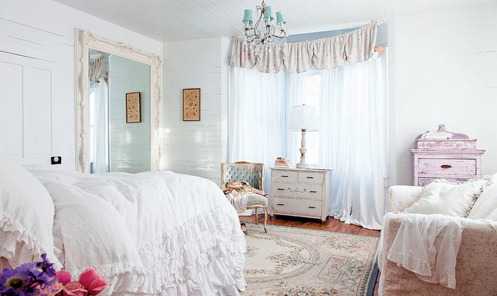 frilly white covers, on a big fluffy bed, near pale patterned, shabby chic sofa, and other vintage furniture 
