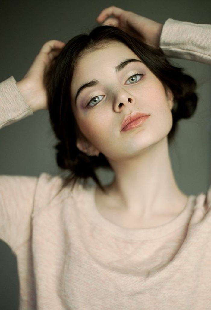 girl with light green eyes, hair parted in the middle, and braided into two side buns, dark brown hair colors, wearing pale pink lipstick, and soft pale pink sweater