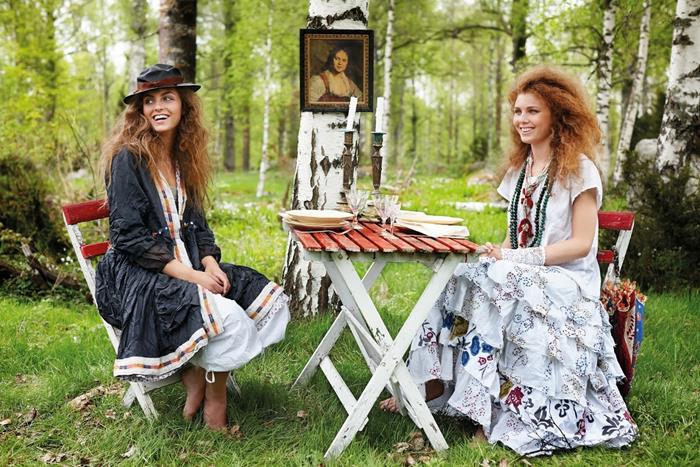 smiling women with voluminous curly hair, sitting on fold-up chairs, near a fold-up table in the woods, wearing bohemian style maxi dresses