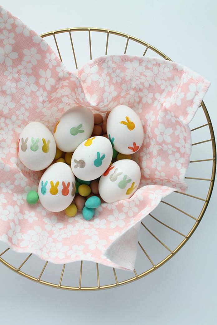 easter egg ideas, pale pink napkin, with white floral pattern, inside a golden basket, containing six white eggs, decorated with multicolored hand-drawn bunny heads, and many small, egg-shaped sweets