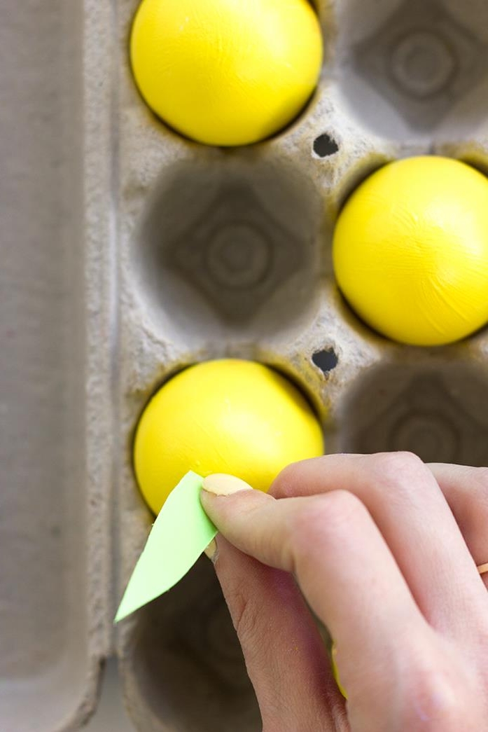 human hand with cream-colored nail polish, sticking a pale grey paper leaf, to a yellow painted egg, dying easter eggs, inside a cardboard box, with two more yellow eggs