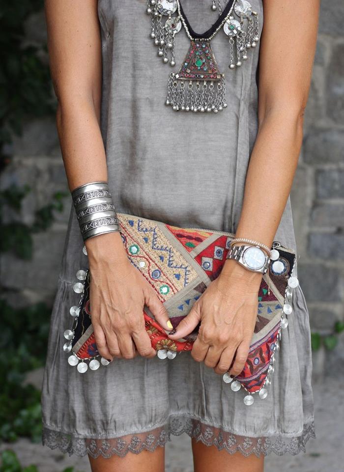 chunky silver tribal necklace with stones, silver bangle and wrist watch, and a multicolored embroidered clutch, with oriental pattern and beads, held by slim woman in grey mini dress, with lace trim, boho style