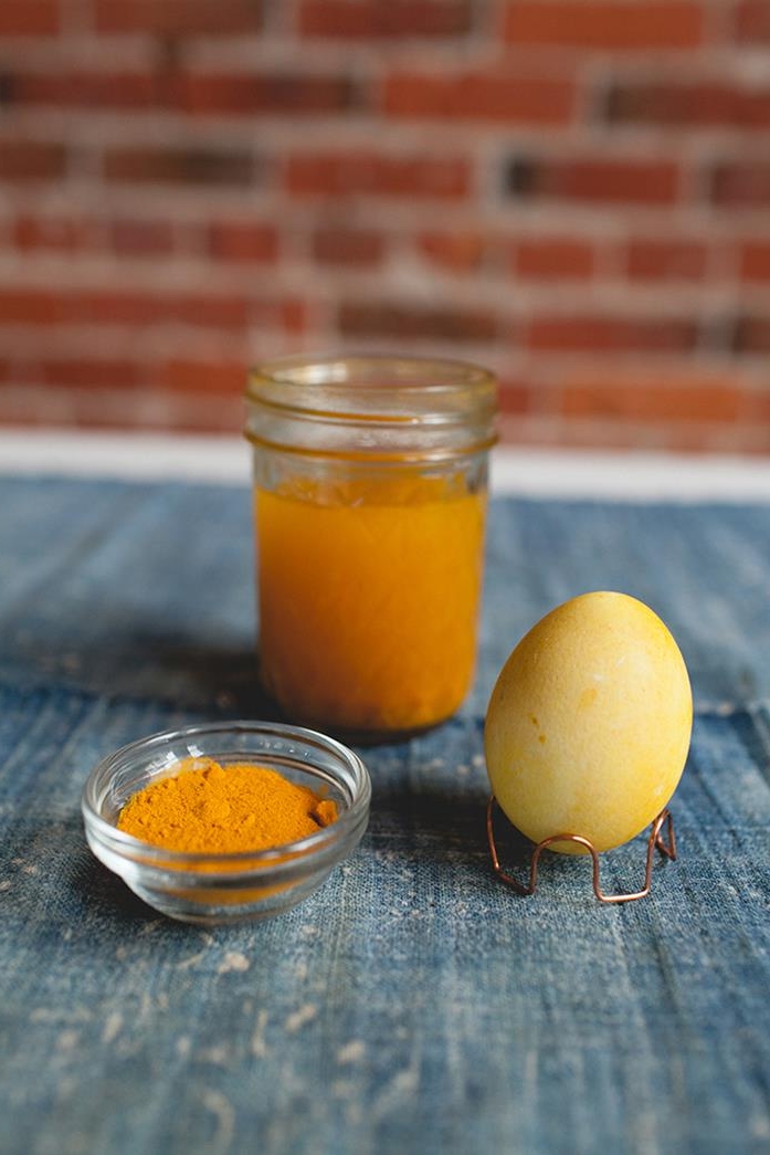 mason jar filled with orange dye, near yellow egg on a wire stand, and a small bowl of ground turmeric, easter egg coloring, with natural ingredients