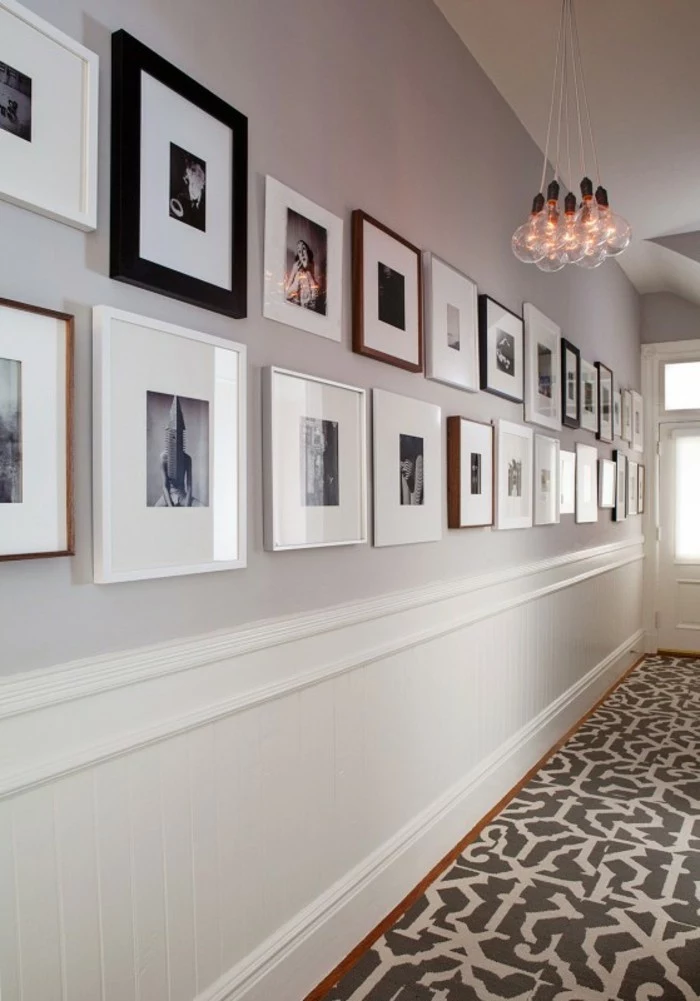 two rows of square frames, in different sizes and colors, containing black and white images, hung on pale gray wall, hallway decor, grey and white floor tiles