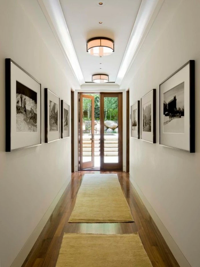 six large square frames, containing black and white images, mounted on two opposite walls of a corridor, hallway decorating ideas, dark wooden floors, two beige rugs