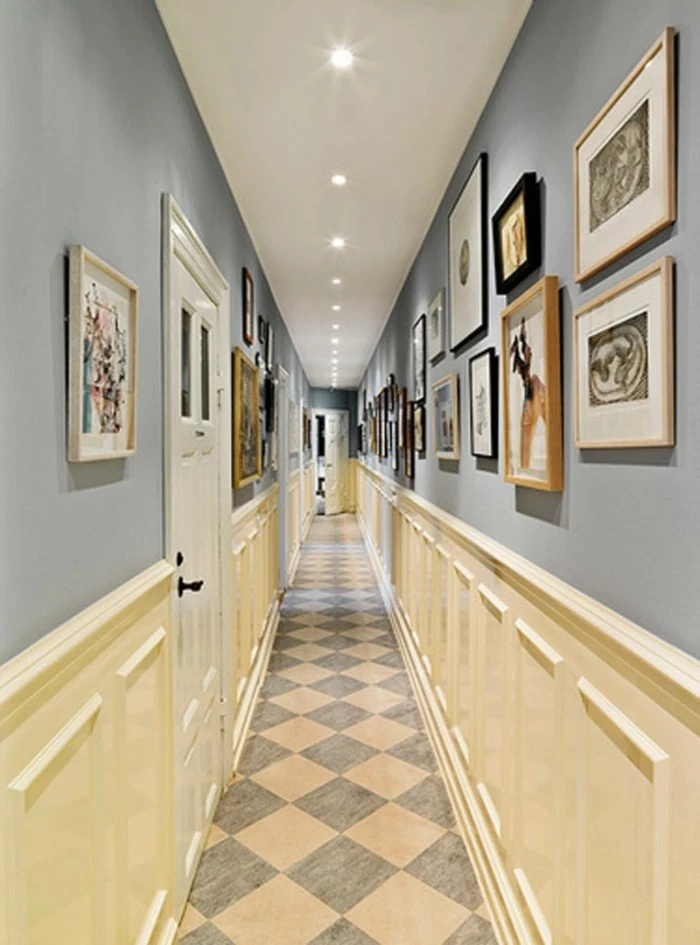 hallway design ideas, long narrow corridor, pale blue-grey walls, with cream white paneling, and many framed images, white ceiling with inbuilt lights, tiled floor and white doors