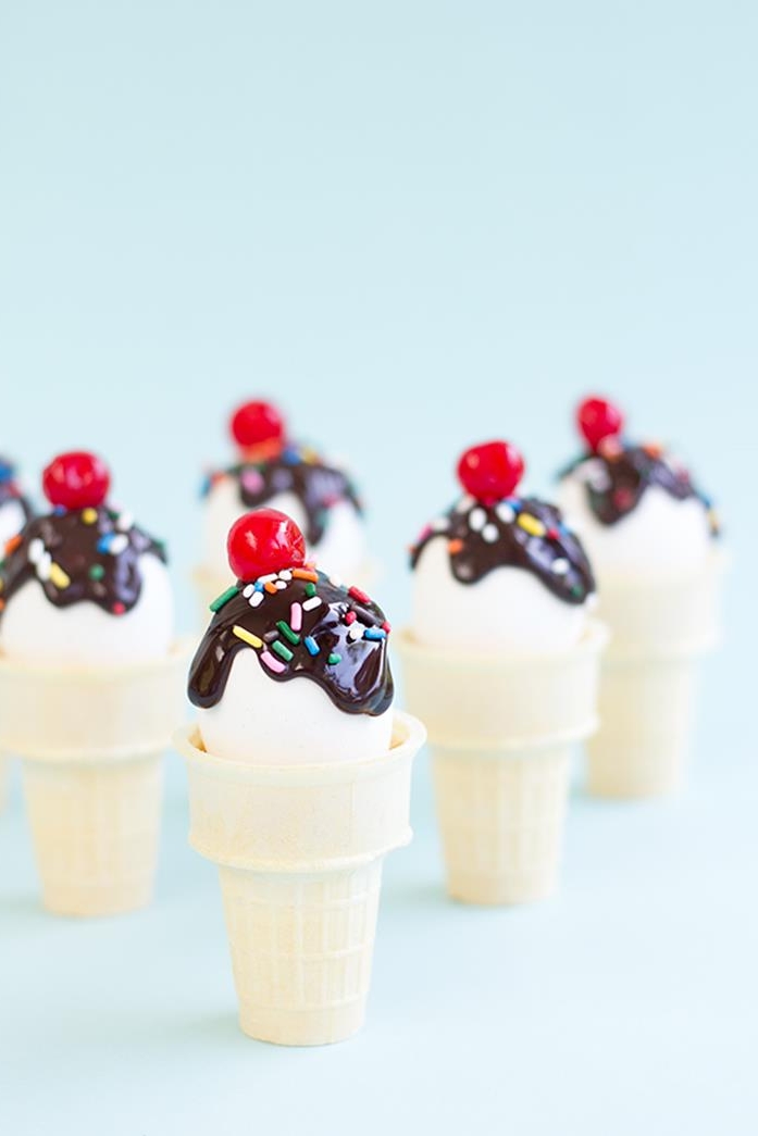 several ice creams, made from white eggs, decorated with brown frosting, multicolored sprinkles, and red cherries, placed inside wafer ice-cream cones, coloring easter eggs, pale blue background