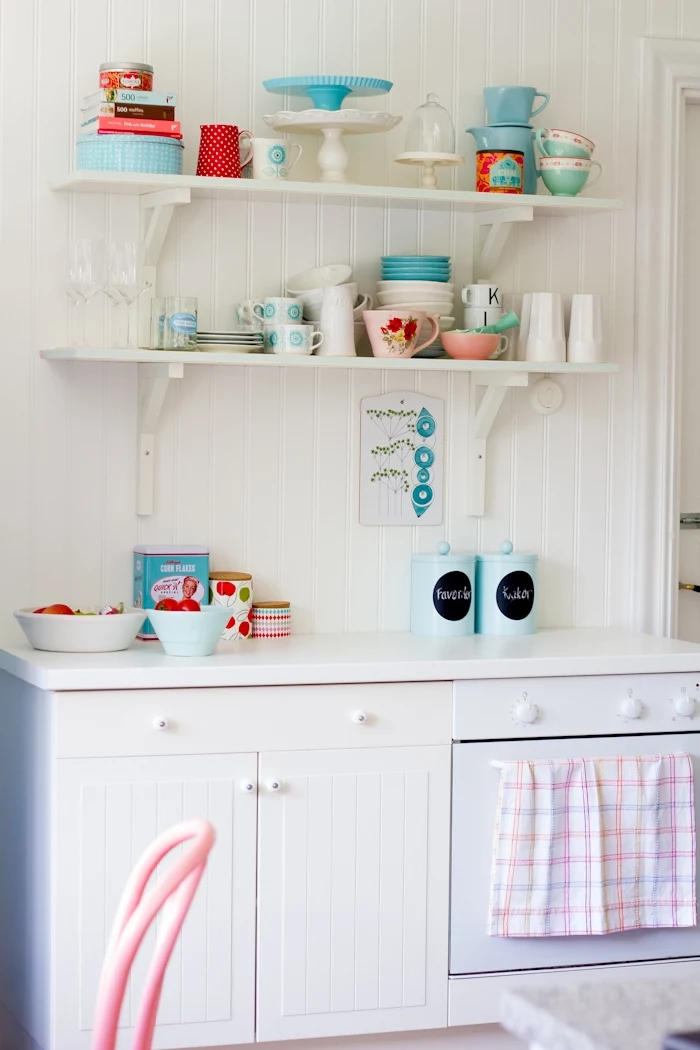 kitchen with white vintage cupboards, two white wooden shelves, shabby chic furniture, various mugs and plates, in pale pastel pink and teal