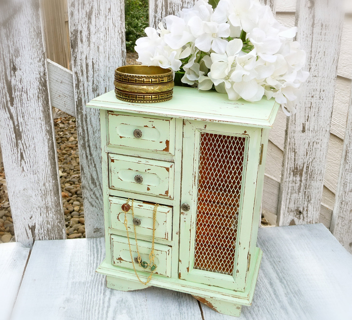 small wooden cabinet in pale green, with four tiny drawers and a compartment, shabby chic furniture, large gold bangle, and white flowers placed on top of it