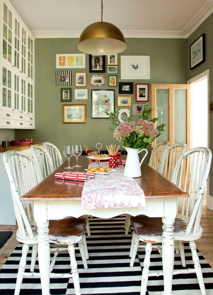 dining room with long wooden table, in white and brown, with matching chairs, shabby chic furniture, green walls with many framed images 