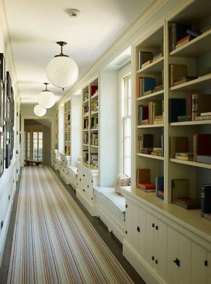 several large white bookshelves, with cupboards underneath, in a long corridor, with white walls, windows and framed images, hallway design ideas, long multicolored striped rug, round ceiling lamps