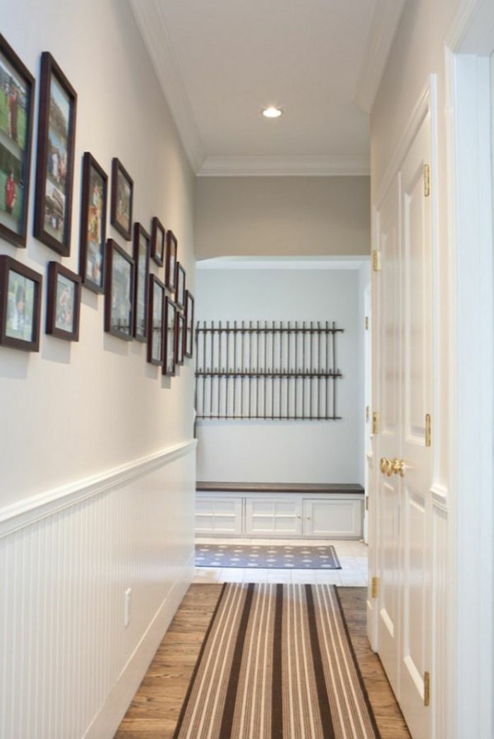 brown frames in different sizes and shapes, containing family photographs, mounted on white wall, small hallway ideas, in corridor with white doors, wooden floor and a striped rug