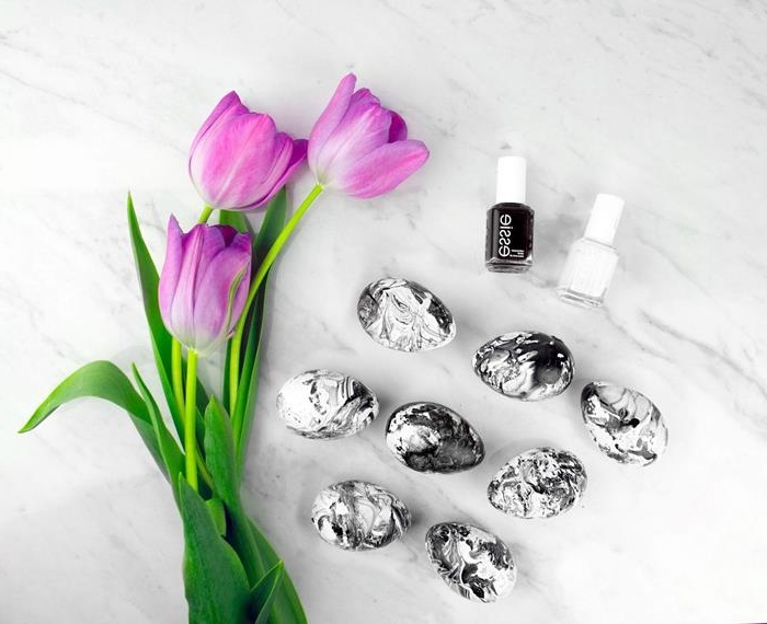 black and white marble effect easter eggs, dyed with nail polish, placed on a marble surface, near three purple tulips, and two bottles of nail polish