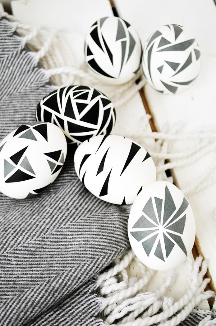 six white eggs, decorated with black and grey triangular shapes, placed on patterned, grey and cream fabric with tassels, dying easter eggs