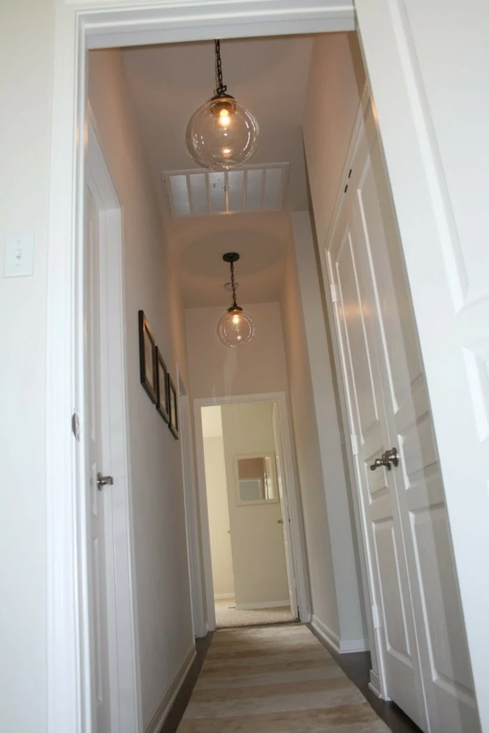 two lit round lamps, made from clear glass, hanging from white ceiling, near two white doors, hallway decor ideas, two white walls, one decorated with three framed images
