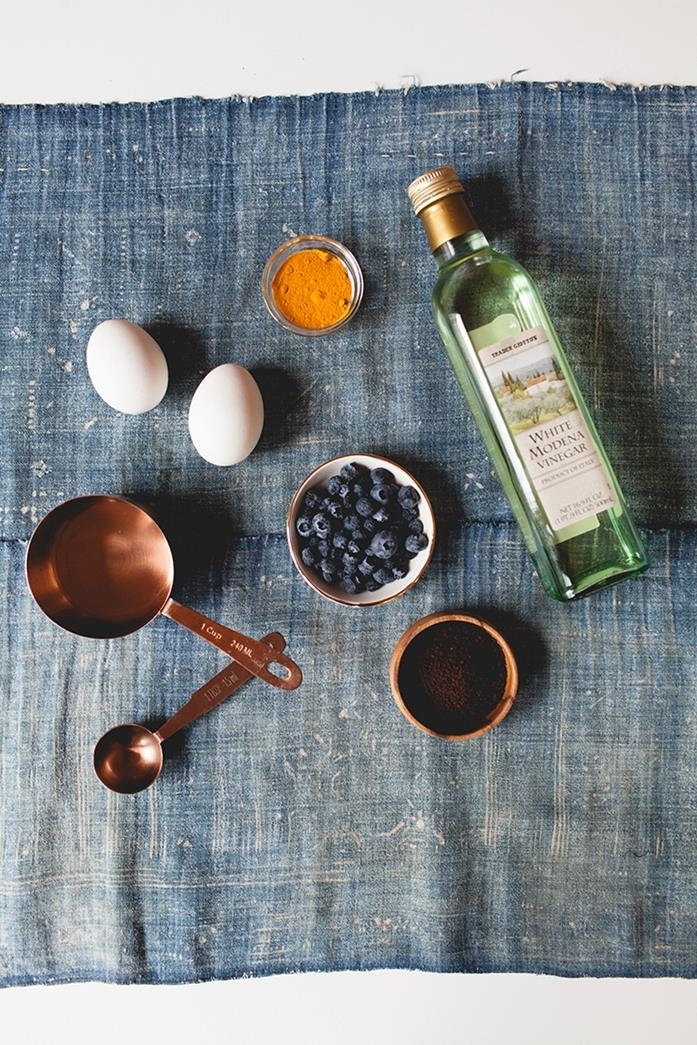 worn blue fabric, with two white eggs, a bottle of vinegar, a bowl of blueberries, a small bowl of ground coffee, a tiny glass bowl of ground turmeric, a small pan and a measuring spoon, easter egg ideas