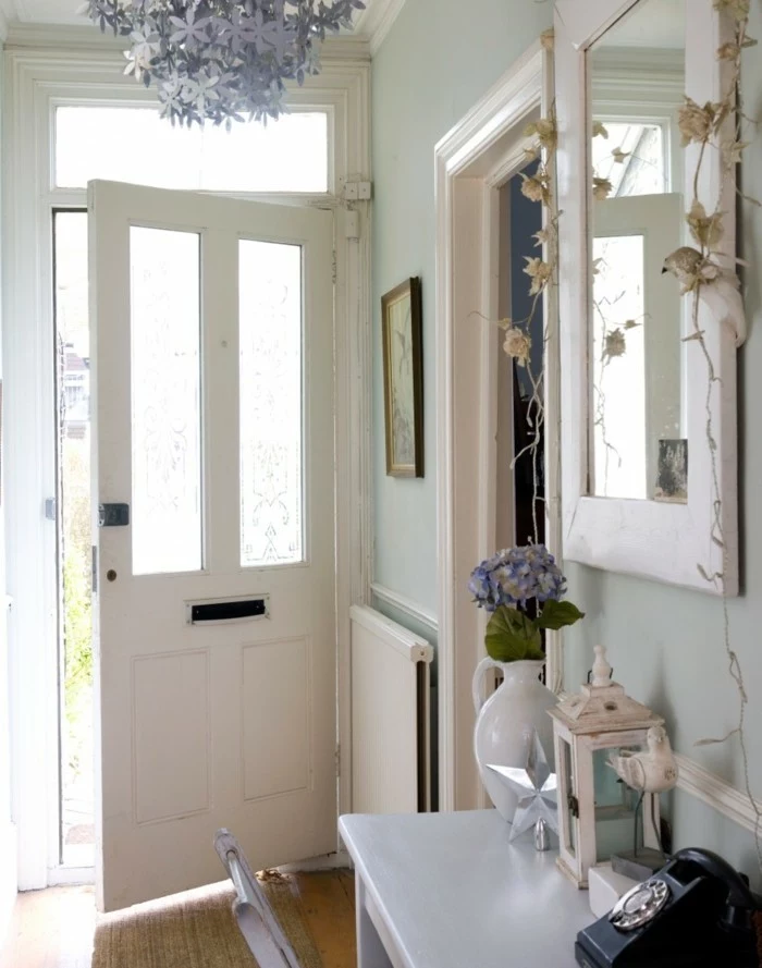 hallway decorating ideas, open white front door, pale blue walls, white plaster details, mirror in white frame, pale grey desk with various items, and matching chair