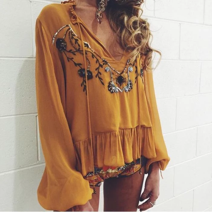 mustard yellow blouse, with long bell sleeves, large frilled hem, and floral embroidery, featuring beads, boho clothing, worn by blonde woman, with curly hair, and multicolored hot pants