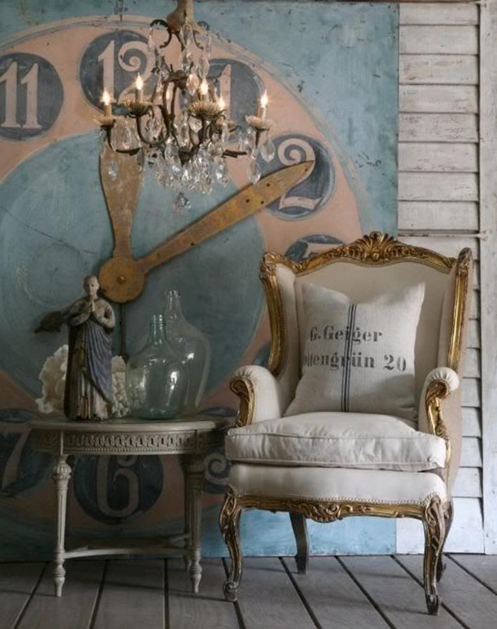 gold and white wing chair, antique shabby chic style, with graphic print cushion, near grey ornamental table, decorated with glass bottles and a statuette, crystal chandelier and a large clock mural