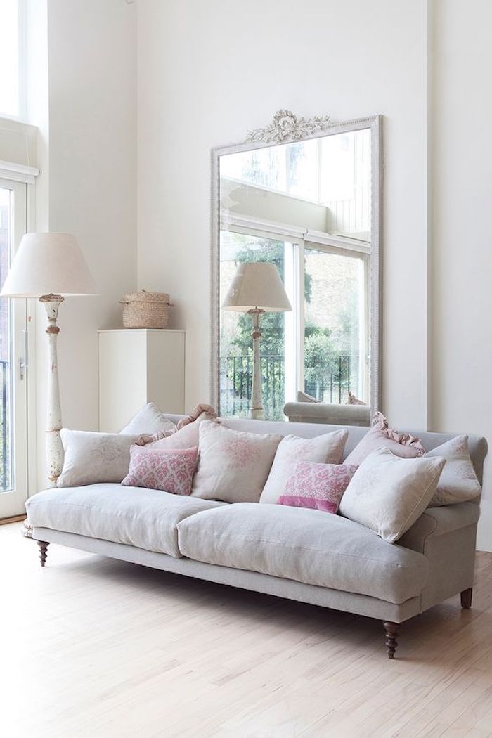 cushions in pale grey and pink, plain and patterned, placed on soft, pale grey modern french sofa, large mirror in white frame, and lamp nearby