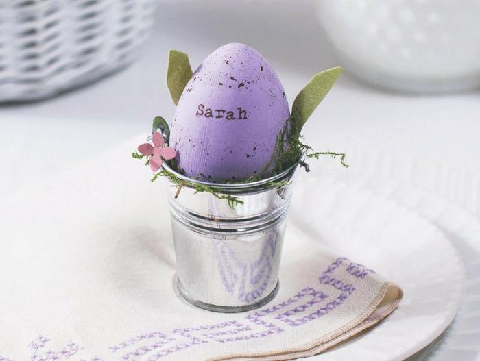 the name sarah, written on a spotty purple egg, placed inside a small metal bucket, decorated with paper flowers and leaves, easter egg decorating, plate with napkin