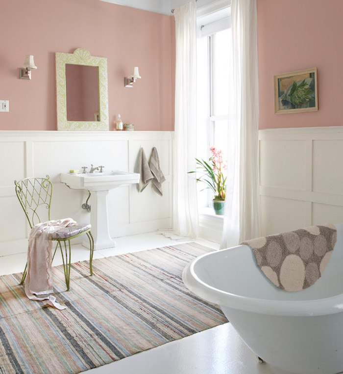 striped multicolored faded rug, in bathroom with pink pastel walls, and white paneling, shabby chic decorating, antique chair and white bathtub, and large window, with sheer cream curtains