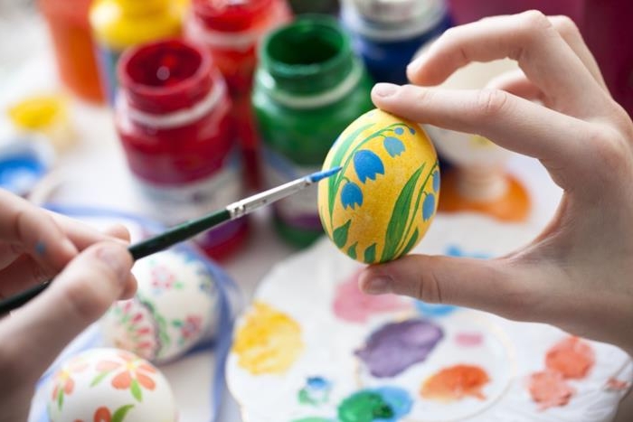 hand holding a painted yellow egg, with blue and green flower, while another hand holds a brush covered in blue near the egg, easter egg designs, paints and palettes in background