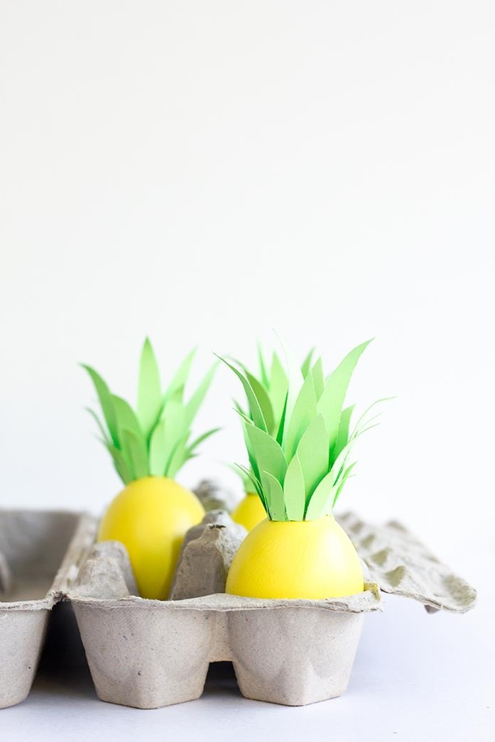 three eggs painted in white, and decorated with many light green paper leaves, to look like pineapples, dying easter eggs, placed inside a grey cardboard egg box