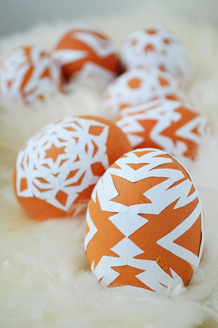 bright orange eggs, decorated with white, symmetrical paper cutouts, forming snowflake-like patterns, easter egg designs, placed on wool-like surface
