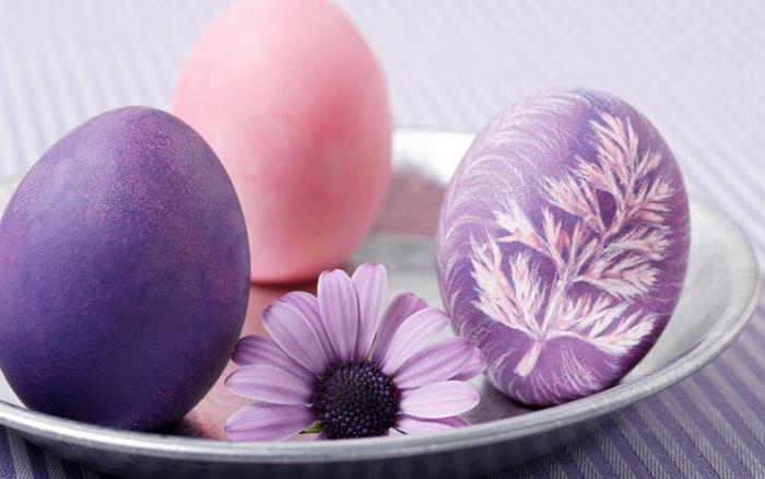 how to dye easter eggs, purple dyed egg, decorated with a crayon drawing of a leaf, in pink and white, placed on a metal plate, near a plain pink and purple egg, and a purple flower