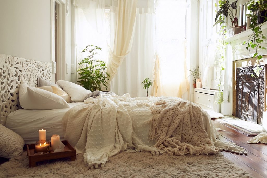 bedroom in white and cream, bed with several blankets pillows and throws, fluffy rug on a wooden floor, tray with lit candles, several different potted plants