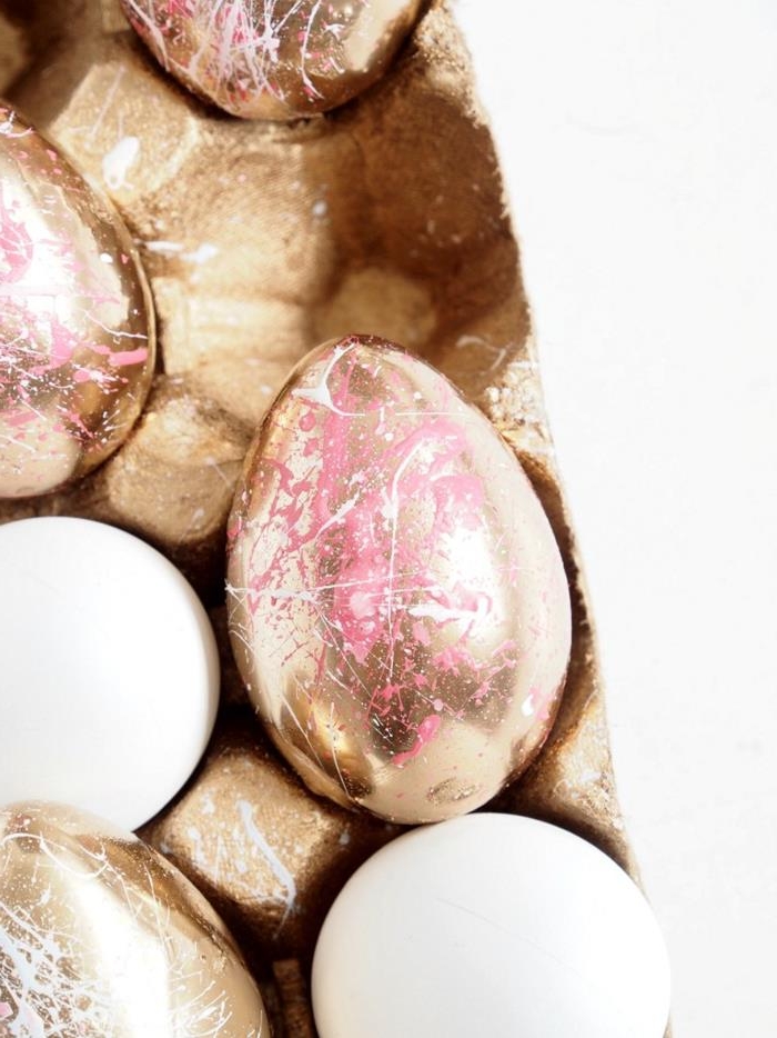 gold painted egg, decorated with drizzles of pink and white paint, easter egg decorating, placed inside a cardboard egg box, near other similar and plain eggs